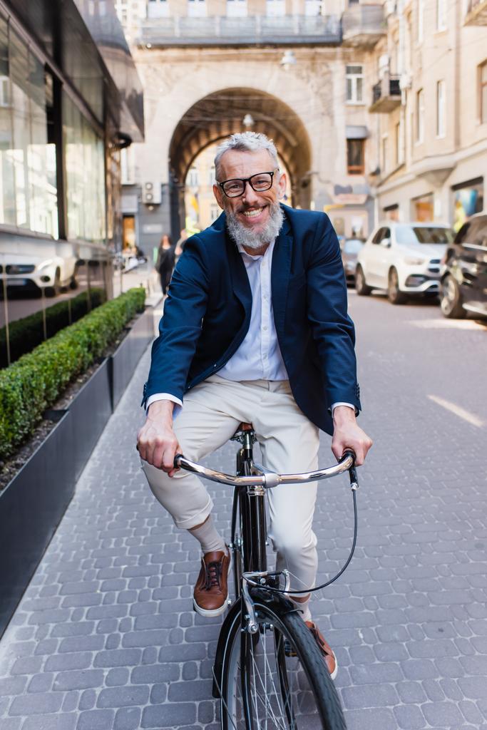 happy middle aged man in glasses riding bicycle on modern urban street  - Photo, Image