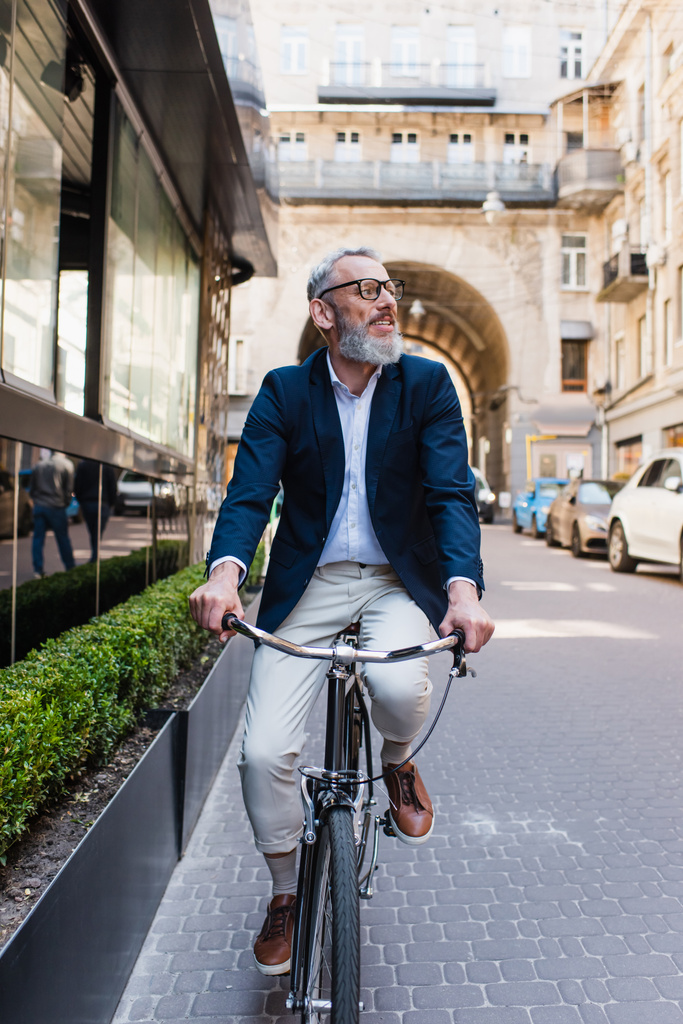 бородатий чоловік середнього віку в окулярах верхи на велосипеді на сучасній міській вулиці
  - Фото, зображення