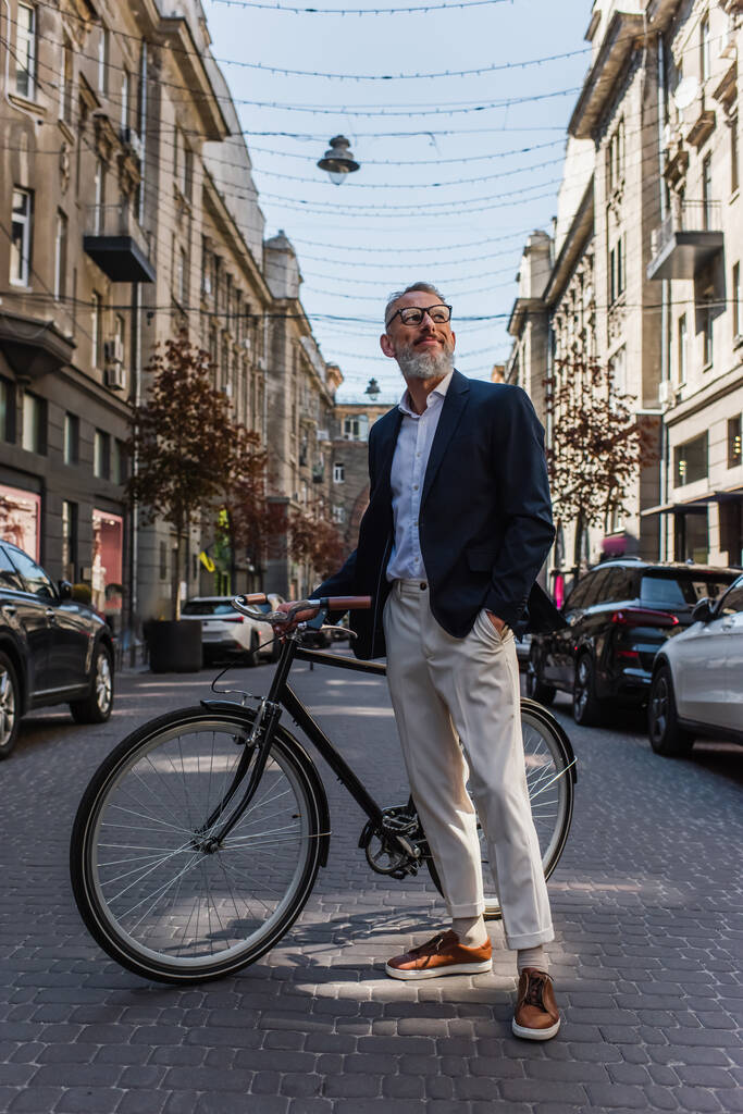 happy middle aged man in blazer and glasses standing with hand in pocket near bicycle on modern urban street  - Zdjęcie, obraz