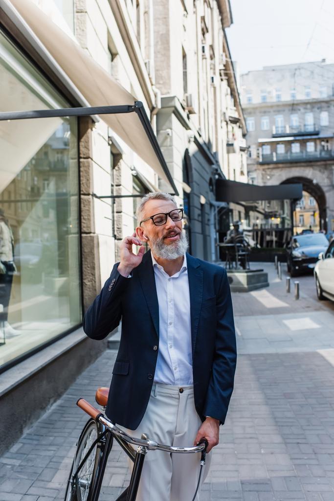 happy mature man in glasses adjusting earphone near bicycle on street  - Valokuva, kuva