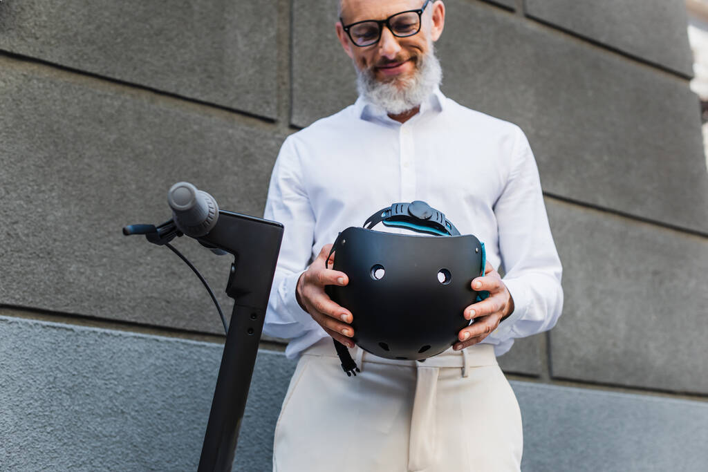 low angle view of smiling and mature man in shirt holding helmet near electric scooter - Zdjęcie, obraz