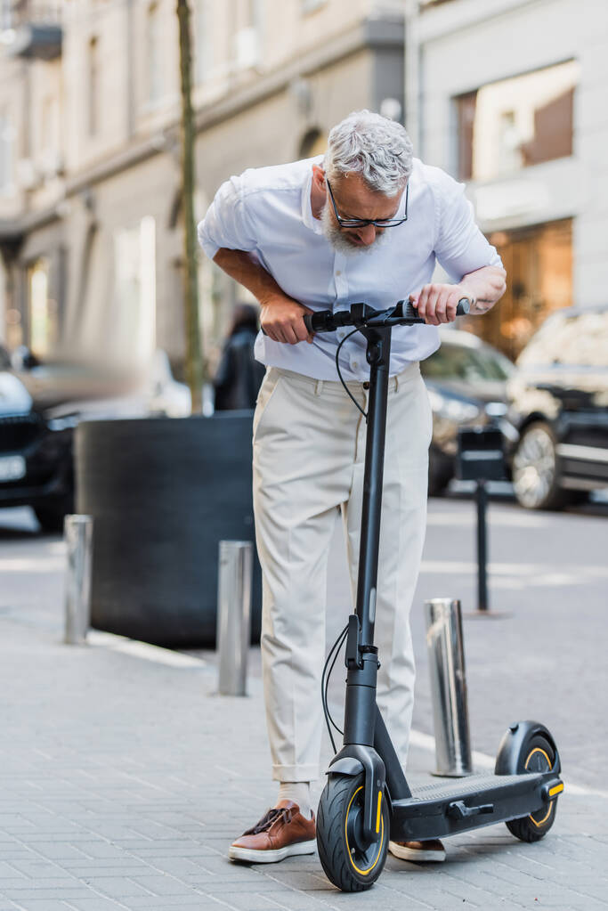 Мужчина в очках в полный рост смотрит на электрический скутер на улице  - Фото, изображение