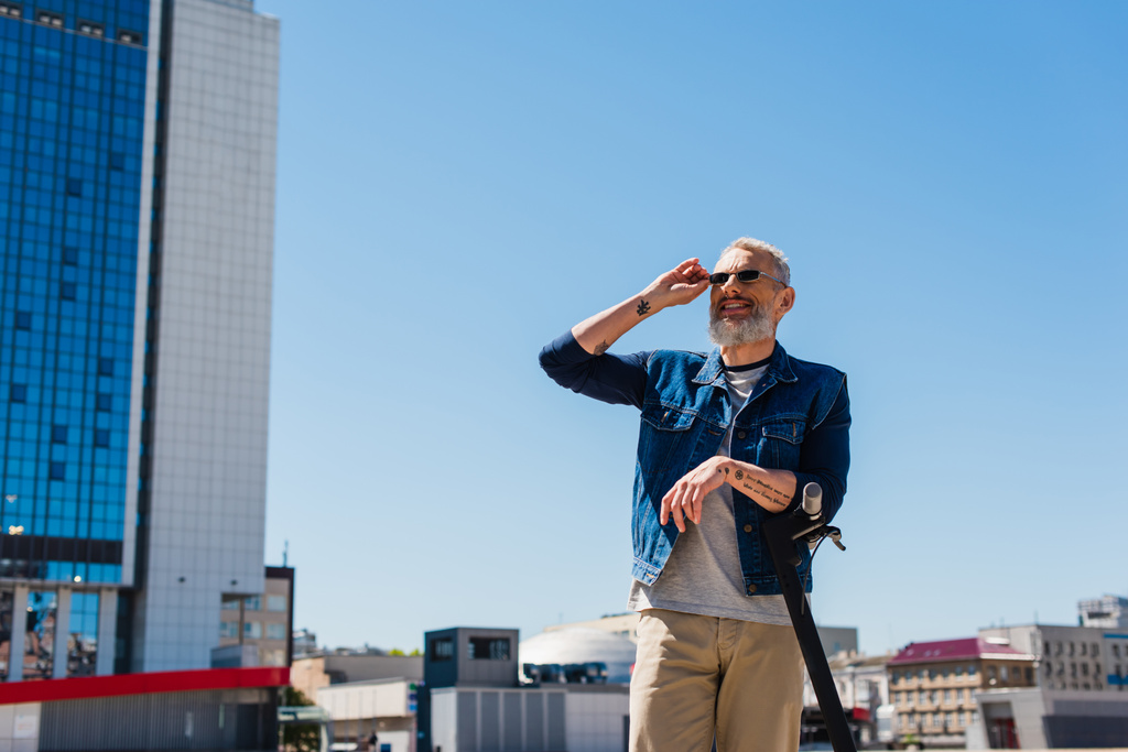 Fröhlicher Mann passt Sonnenbrille neben E-Scooter in Stadtstraße auf - Foto, Bild