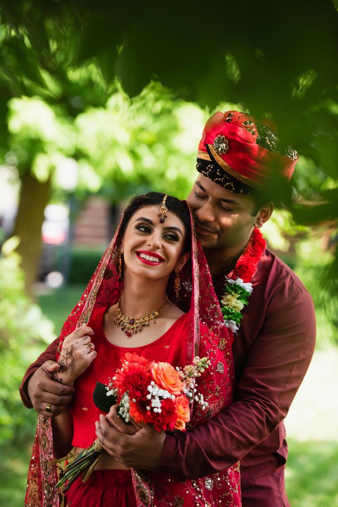 feliz indio hombre abrazando alegre novia en la cabeza bufanda y sari al aire libre - Foto, Imagen