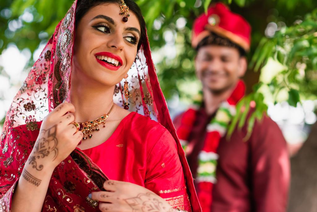 счастливая индийская невеста в сари и головной платок рядом размытый человек в тюрбане на заднем плане  - Фото, изображение