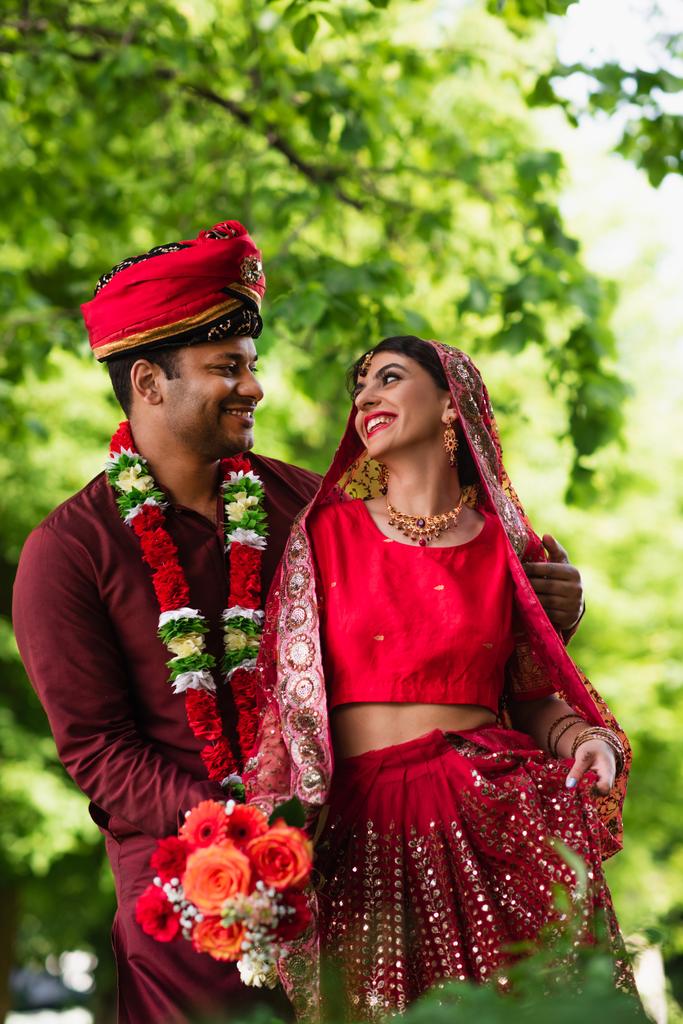 hombre indio positivo en turbante abrazando novia en sari rojo  - Foto, Imagen