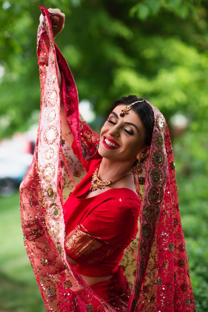 noiva indiana alegre em sari vermelho segurando lenço de cabeça tradicional com ornamento  - Foto, Imagem
