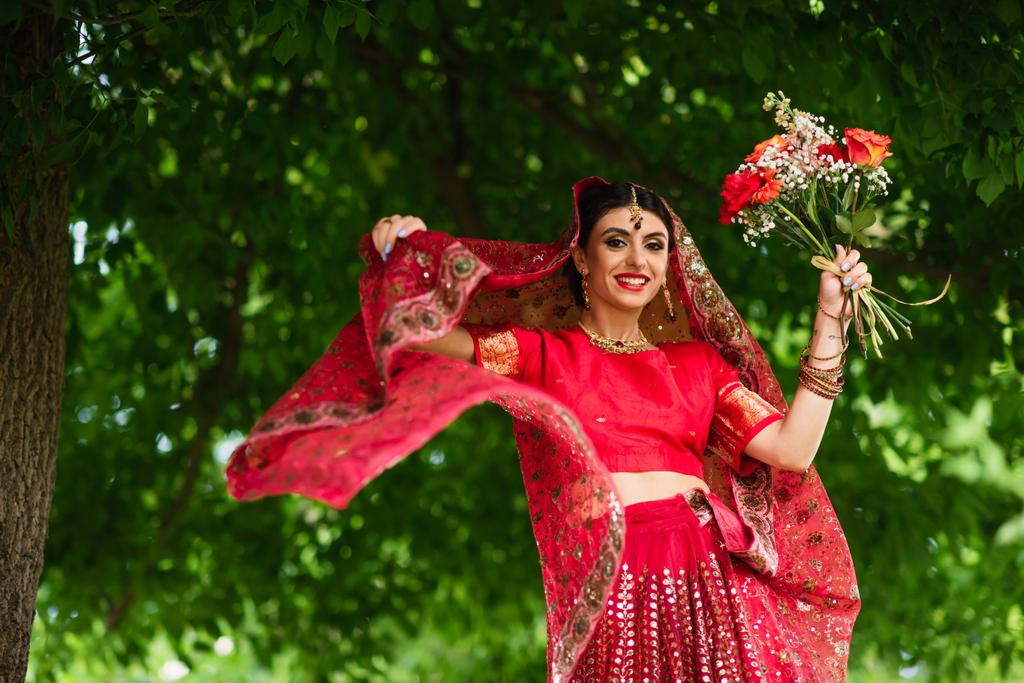 щаслива індійська наречена в червоному сарі і традиційний хустку з орнаментом, що тримає квіти
 - Фото, зображення