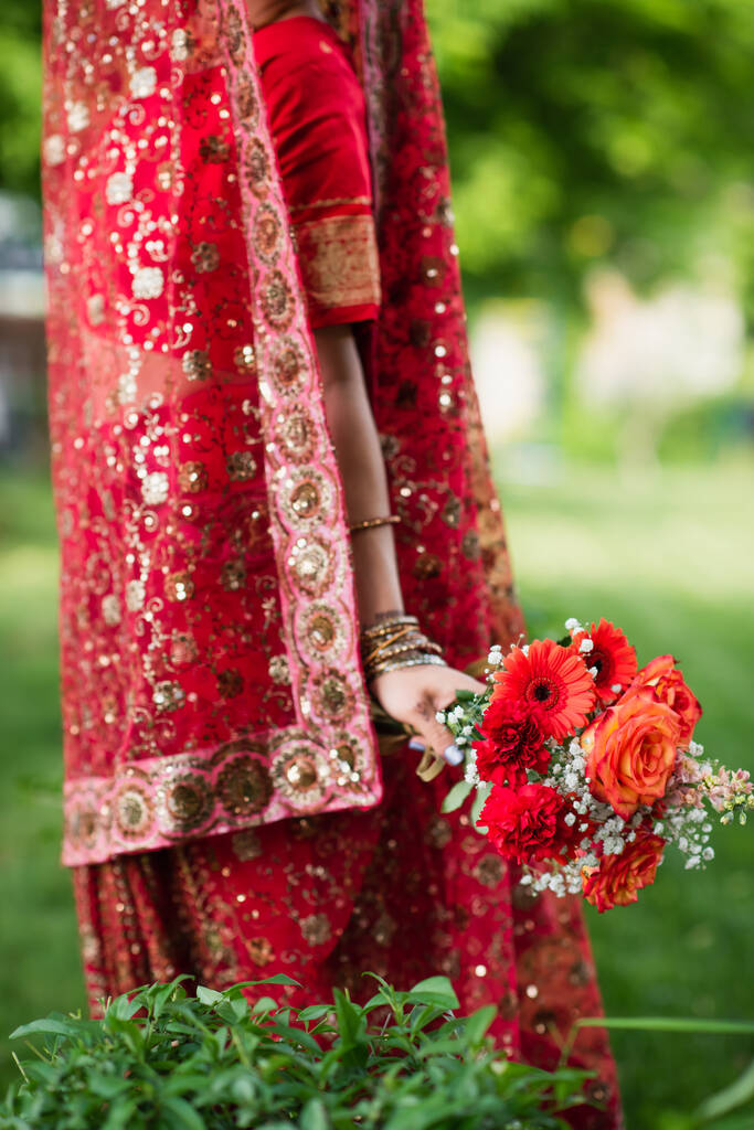 vista recortada de la novia india en sari rojo y pañuelo de cabeza tradicional con adorno sosteniendo flores - Foto, Imagen