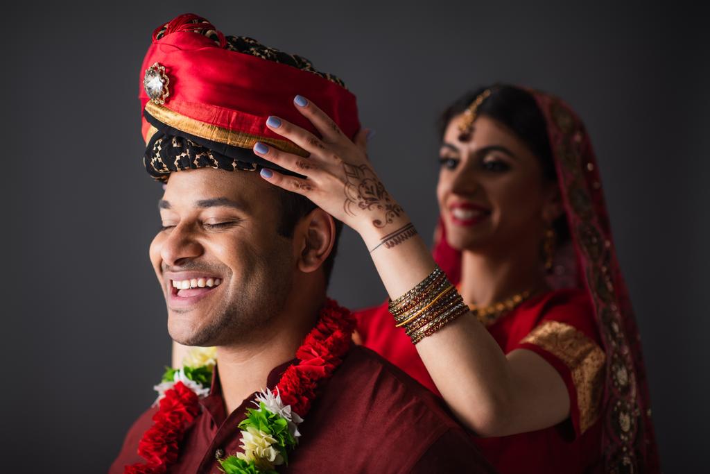 Χαρούμενη Ινδή νύφη που φοράει τουρμπάνι στο κεφάλι του γαμπρού απομονωμένη στο γκρι  - Φωτογραφία, εικόνα