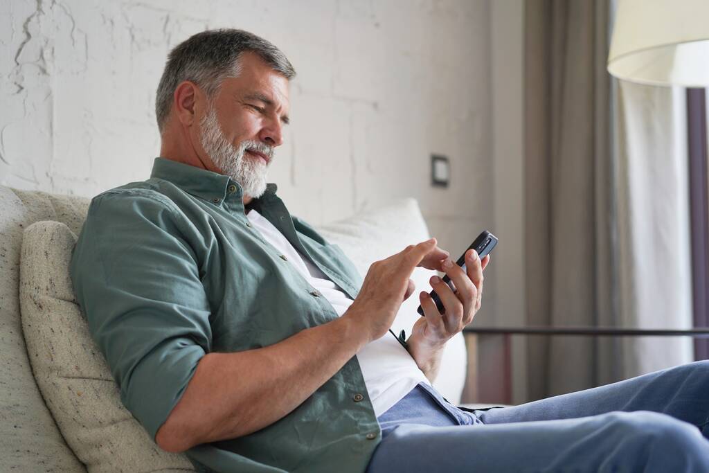 携帯電話を使って笑顔のシニア男性ソファの上に座ってインターネットを閲覧室内. - 写真・画像