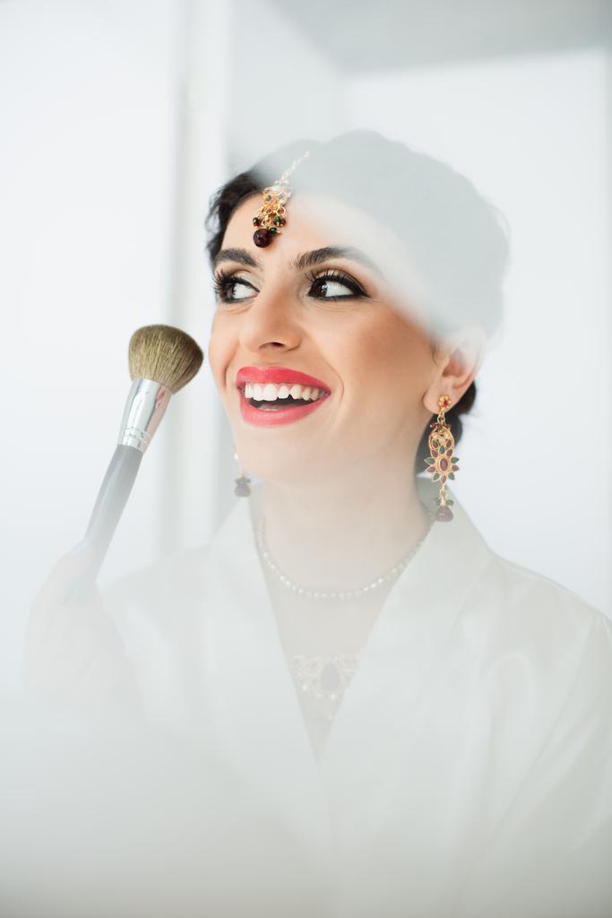 alegre novia india aplicando polvo facial con cepillo cosmético sobre blanco  - Foto, imagen