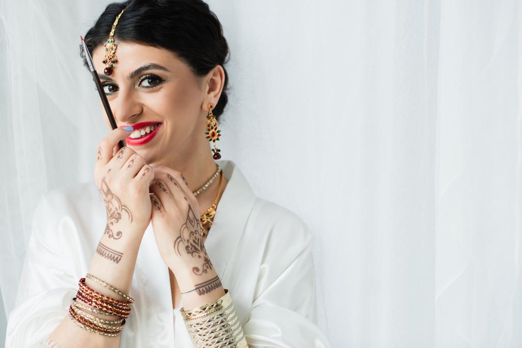 cheerful indian bride with mehndi holding cosmetic brush on white  - Photo, Image