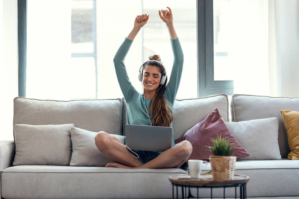 Laukaus motivoitunutta nuorta naista tanssimassa ja kuuntelemassa musiikkia kannettavalla tietokoneellaan istuessaan sohvalla kotona. - Valokuva, kuva