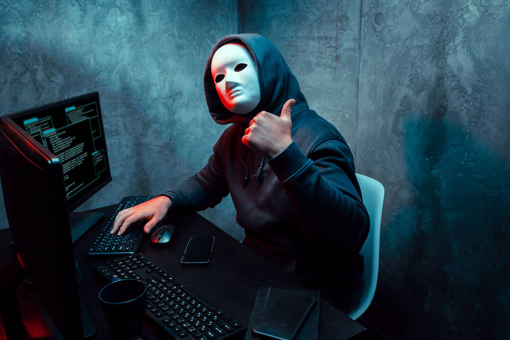 hacker anónimo con máscara facial que trabaja en la computadora en la habitación oscura - Foto, Imagen