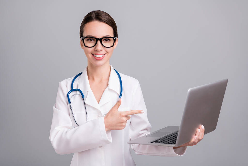 Foto ritratto giovane infermiera con sthethoscope sorridente mostrando dito computer portatile isolato colore grigio sfondo - Foto, immagini