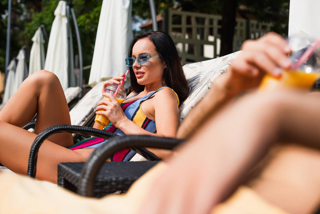 Mulher de roupa de banho segurando suco de laranja na cadeira de praia perto namorado borrado  - Foto, Imagem