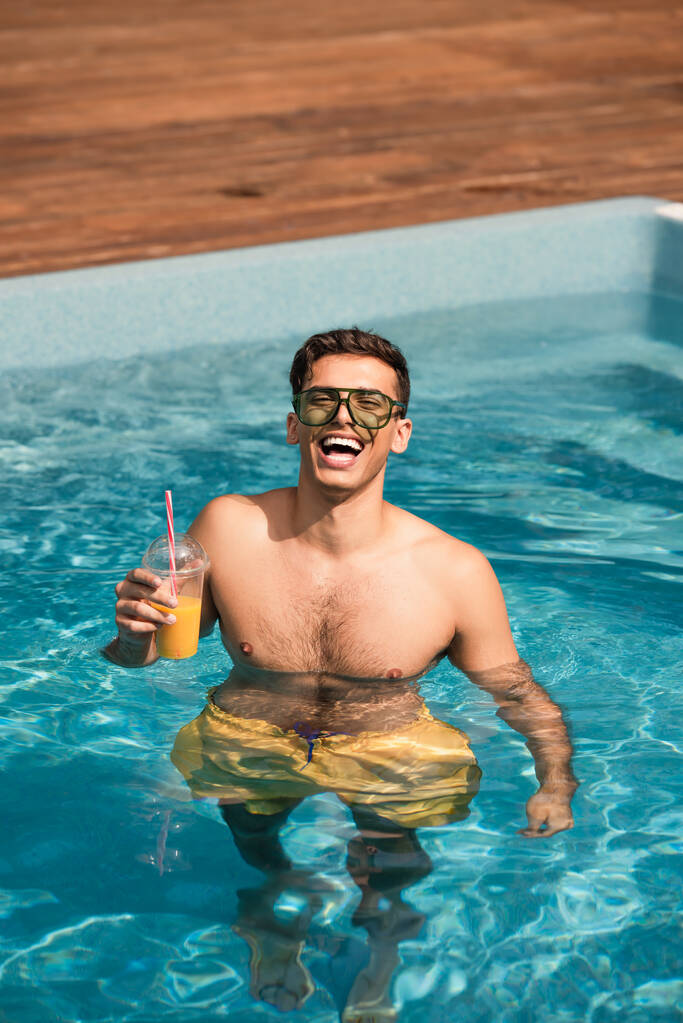 Hombre alegre sosteniendo jugo de naranja en la piscina  - Foto, Imagen