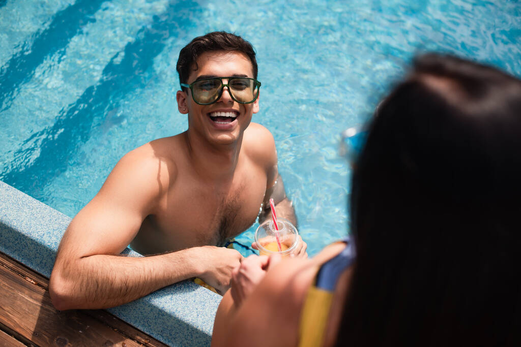 Vista aerea dell'uomo positivo in occhiali da sole che tiene il succo d'arancia vicino alla ragazza offuscata in piscina  - Foto, immagini