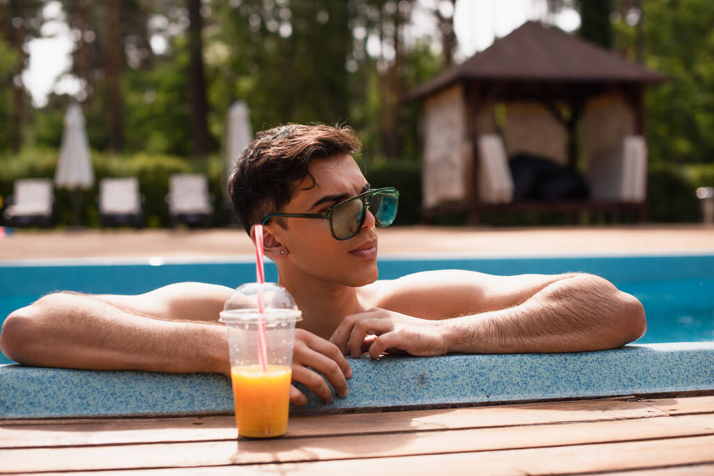 Jeune homme en lunettes de soleil regardant loin près du jus d'orange flou sur le bord de la piscine  - Photo, image