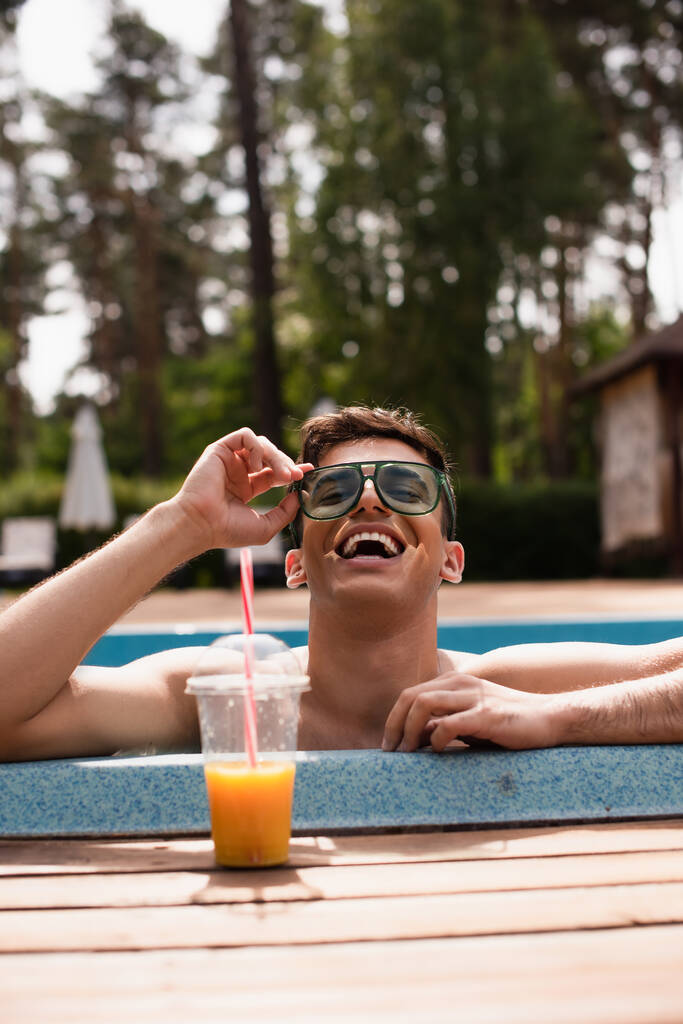 Hombre positivo sosteniendo gafas de sol cerca de jugo de naranja borrosa en la piscina  - Foto, Imagen