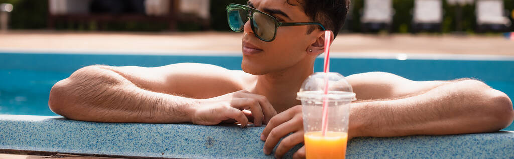 Hombre en gafas de sol relajante en la piscina cerca de jugo de naranja borrosa, pancarta  - Foto, imagen