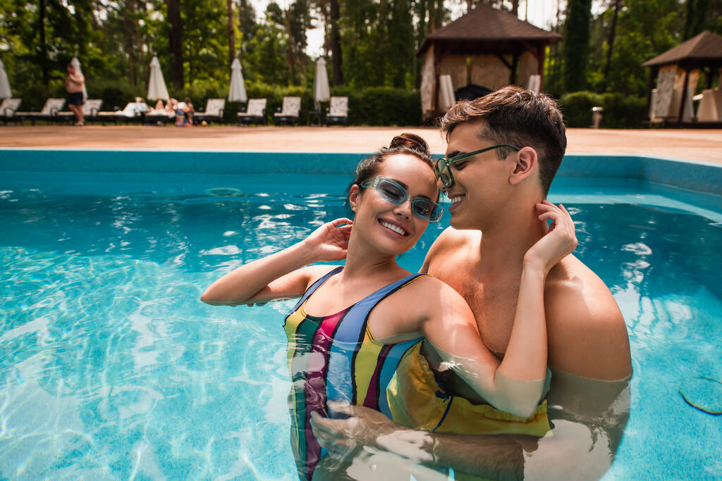 Güneş gözlüklü gülümseyen adam yüzme havuzunda gözleri kapalı kız arkadaşına sarılıyor.  - Fotoğraf, Görsel