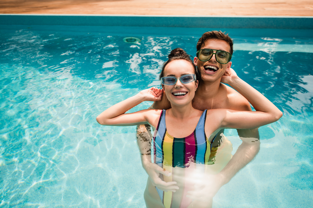 Boldog pár napszemüvegben nézi a kamerát, miközben a medencében állnak.  - Fotó, kép