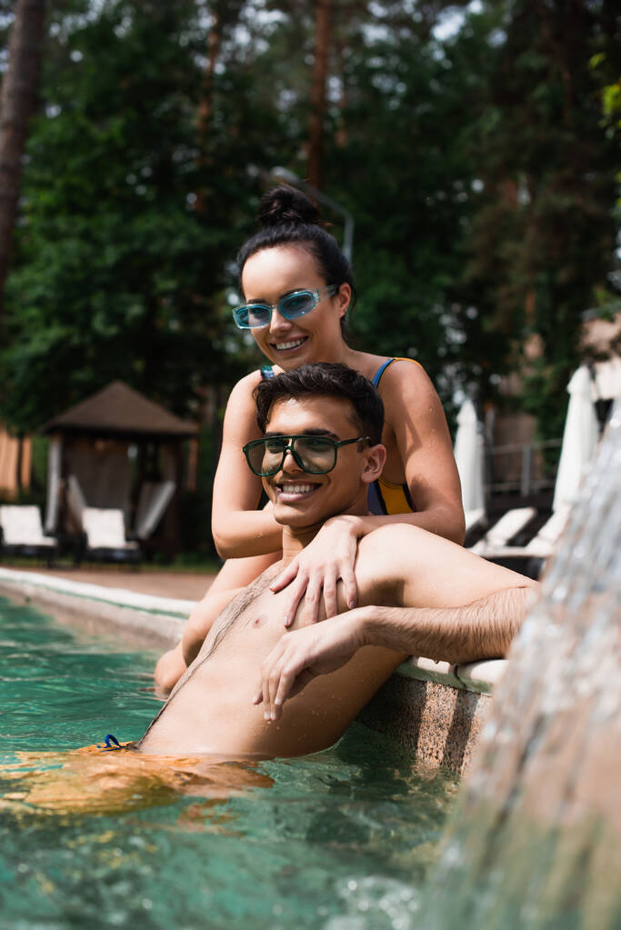 Smiling woman relaxing with boyfriend in swimming pool near blurred fountain  - Photo, Image