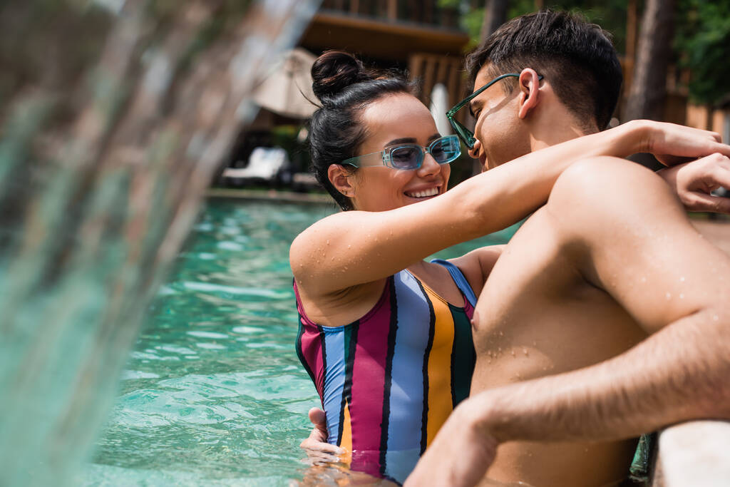 Молода пара в сонцезахисних окулярах обіймається біля розмитого фонтану в басейні
  - Фото, зображення
