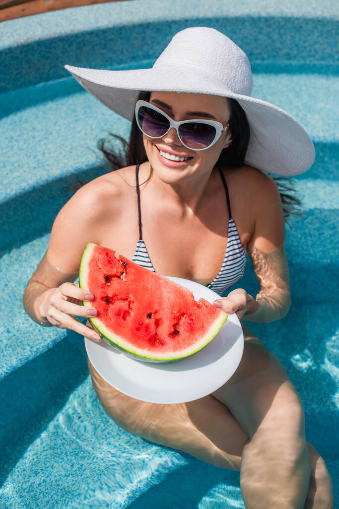 Vista de alto ângulo da mulher positiva em roupa de banho segurando melancia na piscina  - Foto, Imagem