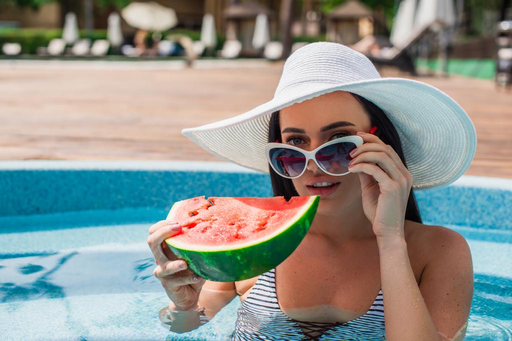 Женщина в солнечных очках держит арбуз и смотрит на камеру в бассейне  - Фото, изображение
