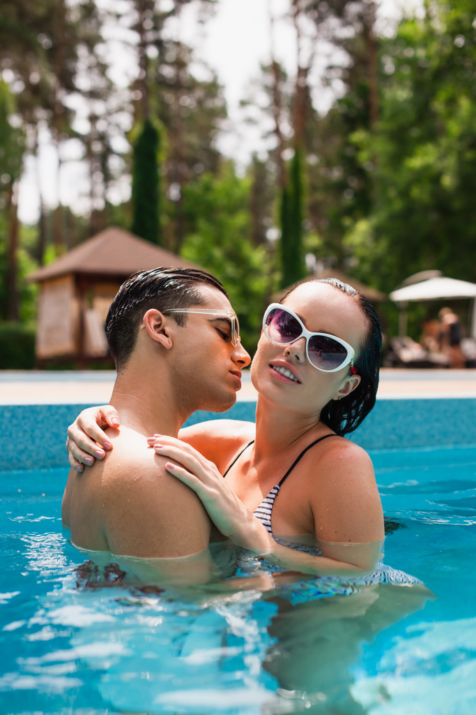 Мужчина целует красивую девушку в солнечных очках в бассейне  - Фото, изображение