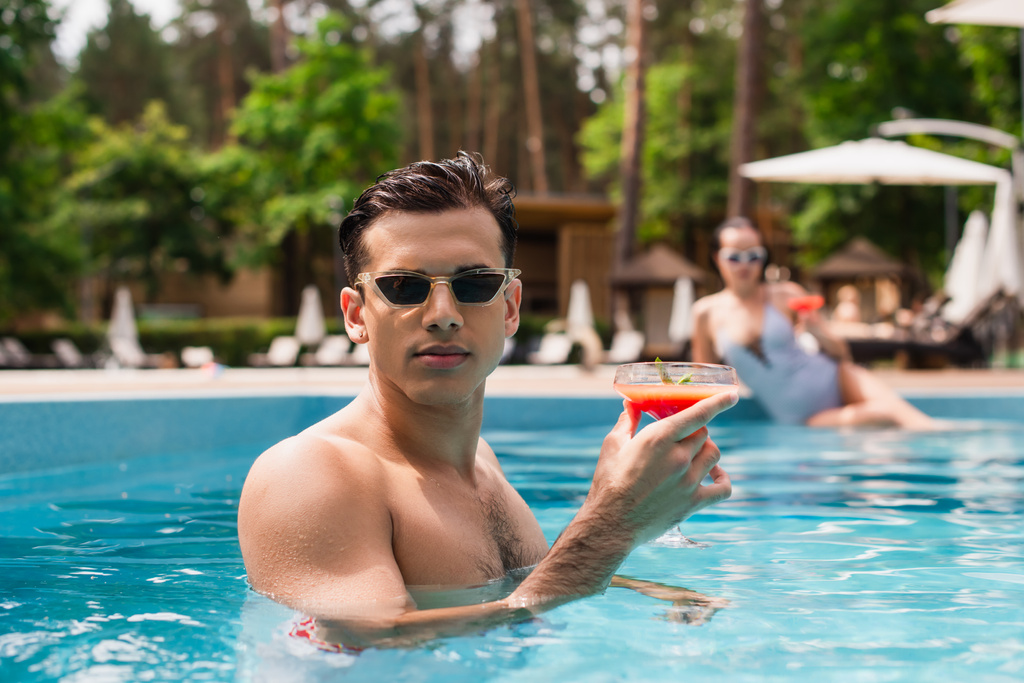 Young man holding cocktail near blurred girlfriend in swimming pool on blurred background  - 写真・画像