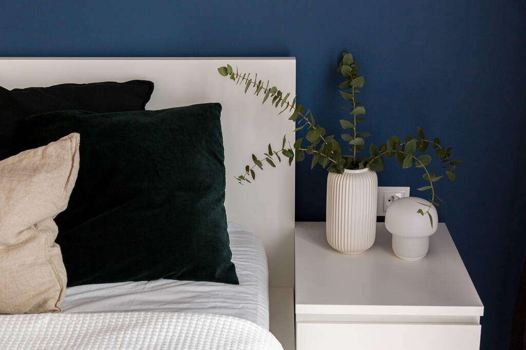 Weißer Nachttisch mit Zierlampe und Vase mit Eukalyptuszweigen neben gemütlichem Bett im Schlafzimmer mit marineblauer Wand - Foto, Bild