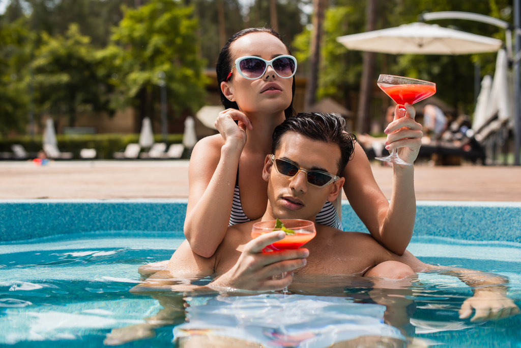Брюнетка держит коктейль, расслабляясь рядом с парнем в бассейне  - Фото, изображение