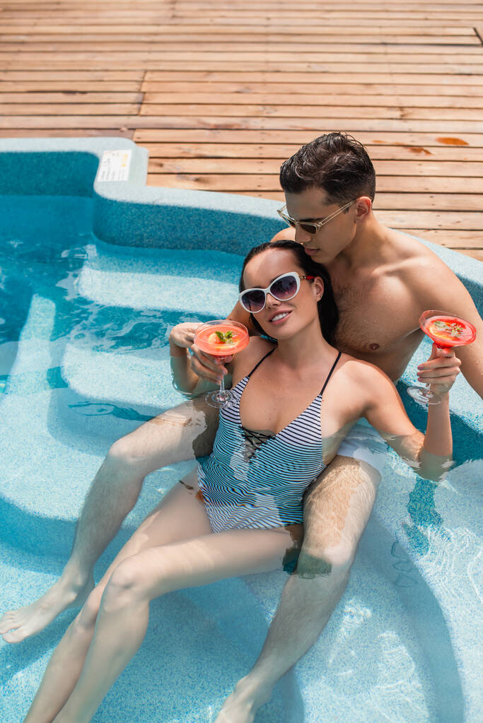 Wysoki kąt widzenia uśmiechnięta kobieta trzyma koktajl w pobliżu chłopaka w okularach przeciwsłonecznych w basenie  - Zdjęcie, obraz
