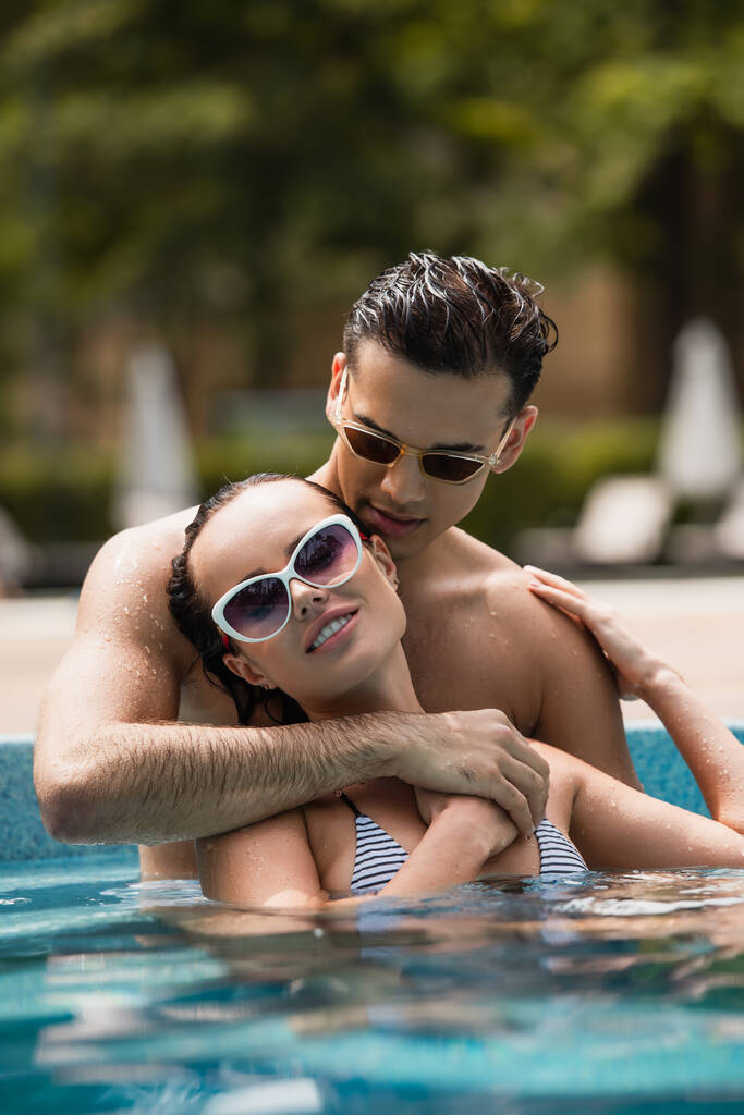 Yüzme havuzunda, güneş gözlüklü, gülümseyen kadını kucaklayan adamın yüzey seviyesi.  - Fotoğraf, Görsel