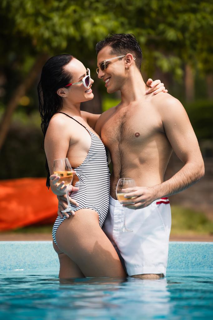 Vista lateral de mujer sonriente con novio abrazador de vino en piscina  - Foto, Imagen