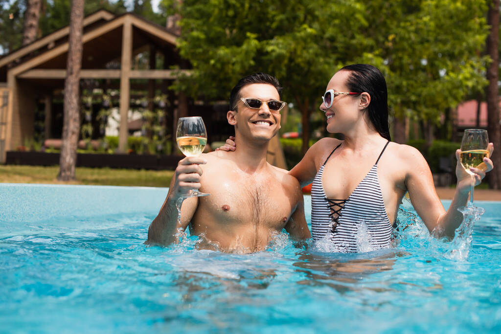 Ζευγάρι με ποτήρια κρασιού χαμογελώντας στην εξωτερική πισίνα  - Φωτογραφία, εικόνα