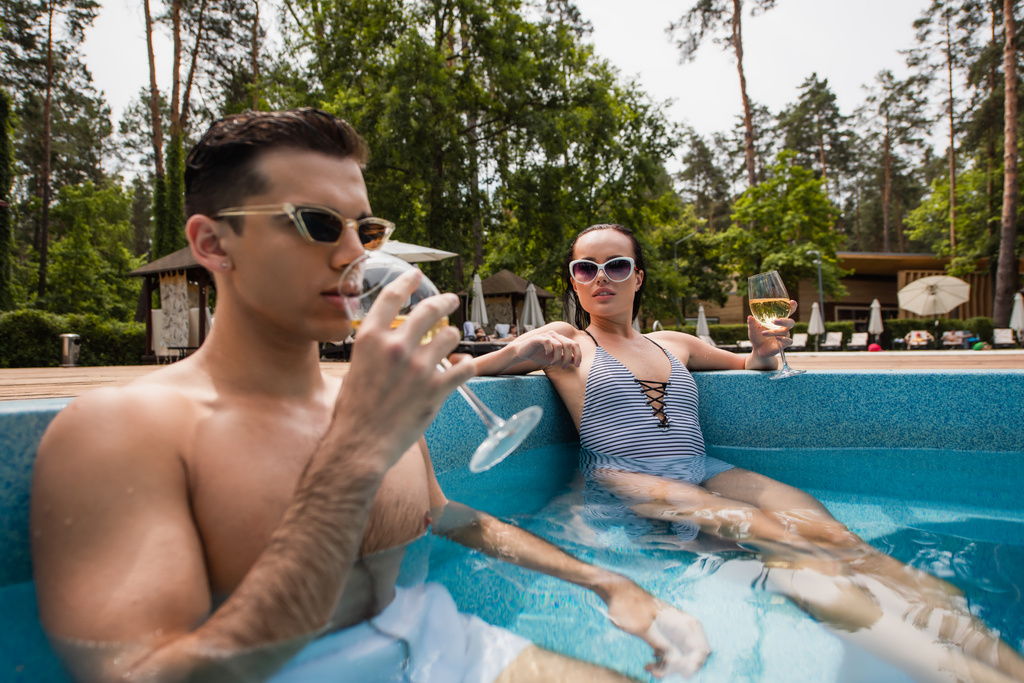 Junge Frau in gestreifter Badebekleidung sitzt neben Freund und trinkt Wein im Pool  - Foto, Bild