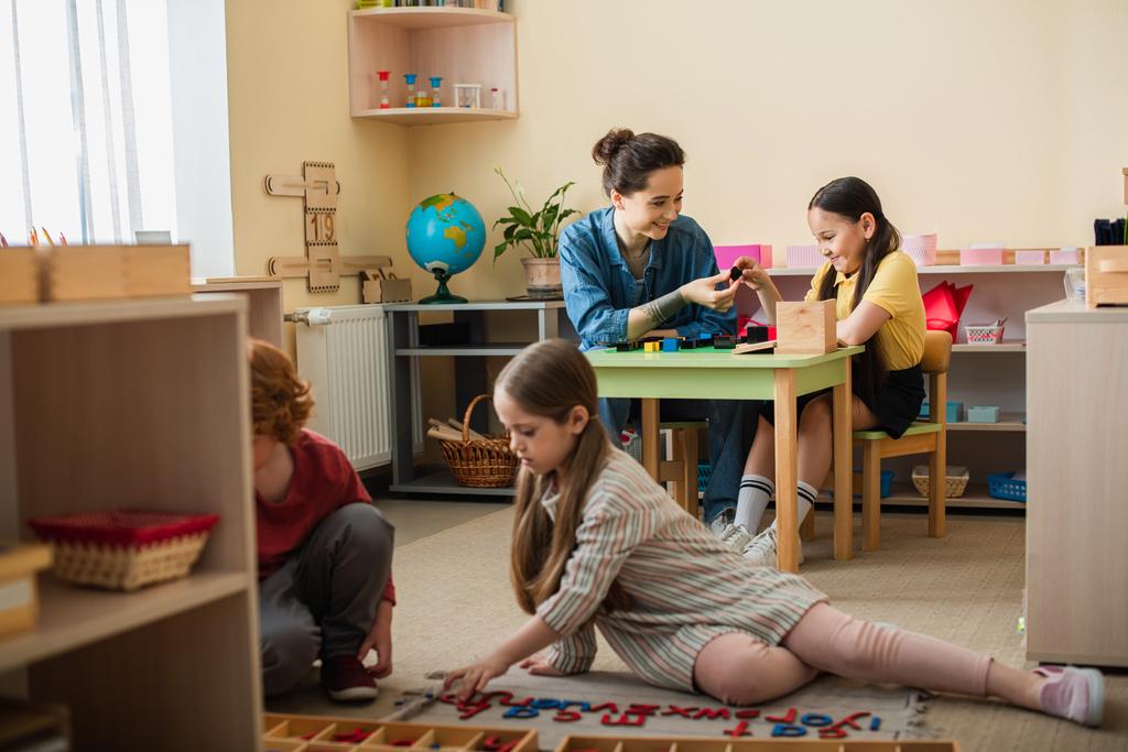 enfants sur le sol jouer avec des lettres en bois près de professeur et fille avec des cubes multicolores sur fond - Photo, image