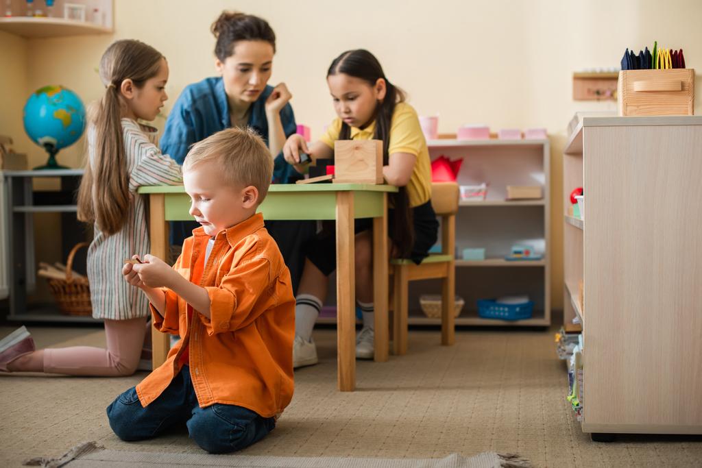 хлопчик грає на підлозі біля міжрасових дівчат і вчителька на розмитому фоні
 - Фото, зображення