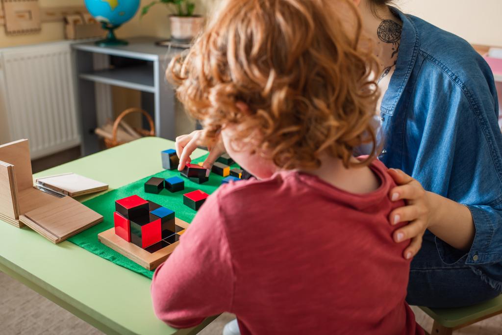 πίσω όψη της κοκκινομάλλα αγόρι παίζει εκπαιδευτικό παιχνίδι με το δάσκαλο στο Montessori σχολείο - Φωτογραφία, εικόνα