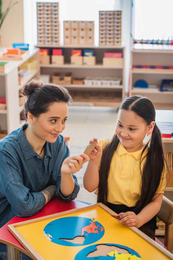 joven profesor dando pieza de rompecabezas a asiático chica en montessori escuela - Foto, Imagen