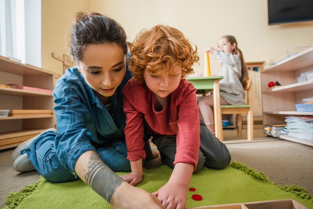 getatoeëerde leraar spelen op de vloer met roodharige jongen in de buurt meisje op wazig achtergrond - Foto, afbeelding