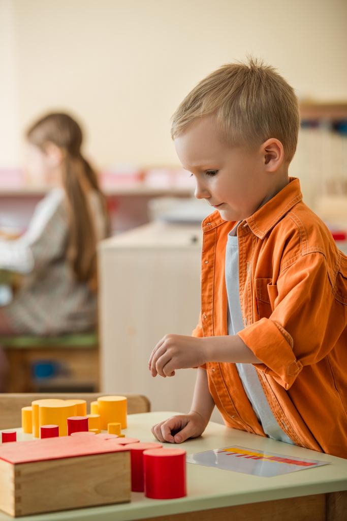 хлопчик грає з червоними і жовтими циліндрами біля розмитої дівчини в школі Монтессорі
 - Фото, зображення
