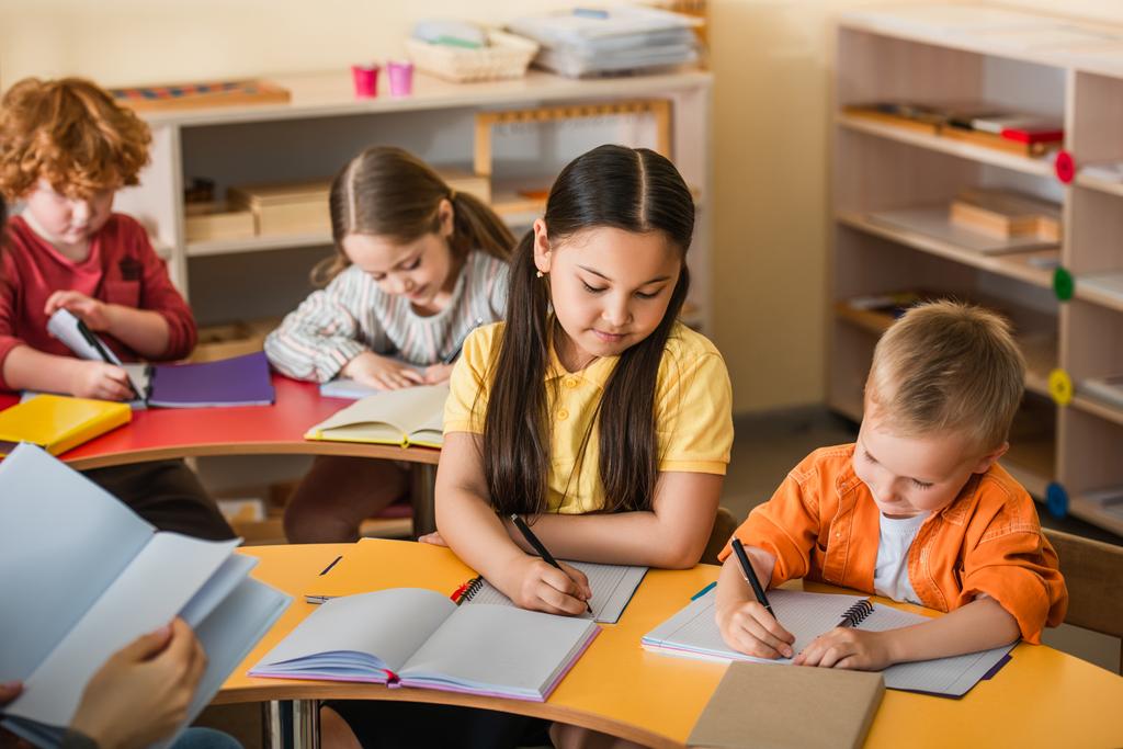 multiethnische Kinder schreiben in Notizbüchern während des Unterrichts in der Montessori-Schule - Foto, Bild