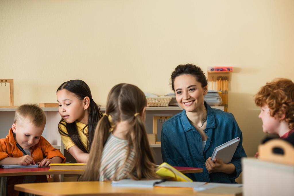 jovem professor com livro sorrindo perto de crianças inter-raciais na sala de aula montessori - Foto, Imagem