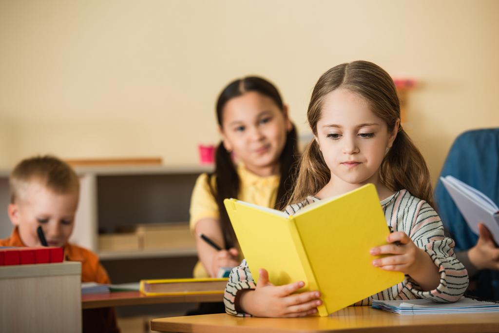 κορίτσι ανάγνωση του βιβλίου κατά τη διάρκεια Leson στο Montessori σχολείο κοντά θολή πολυεθνική φίλους - Φωτογραφία, εικόνα